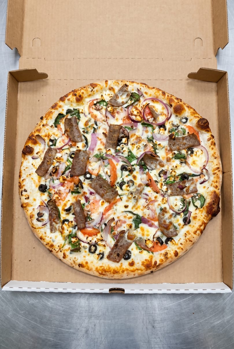 A whole pizza with white sauce, gyro meat, red onions, tomatoes, black olives, and feta cheese in a box.