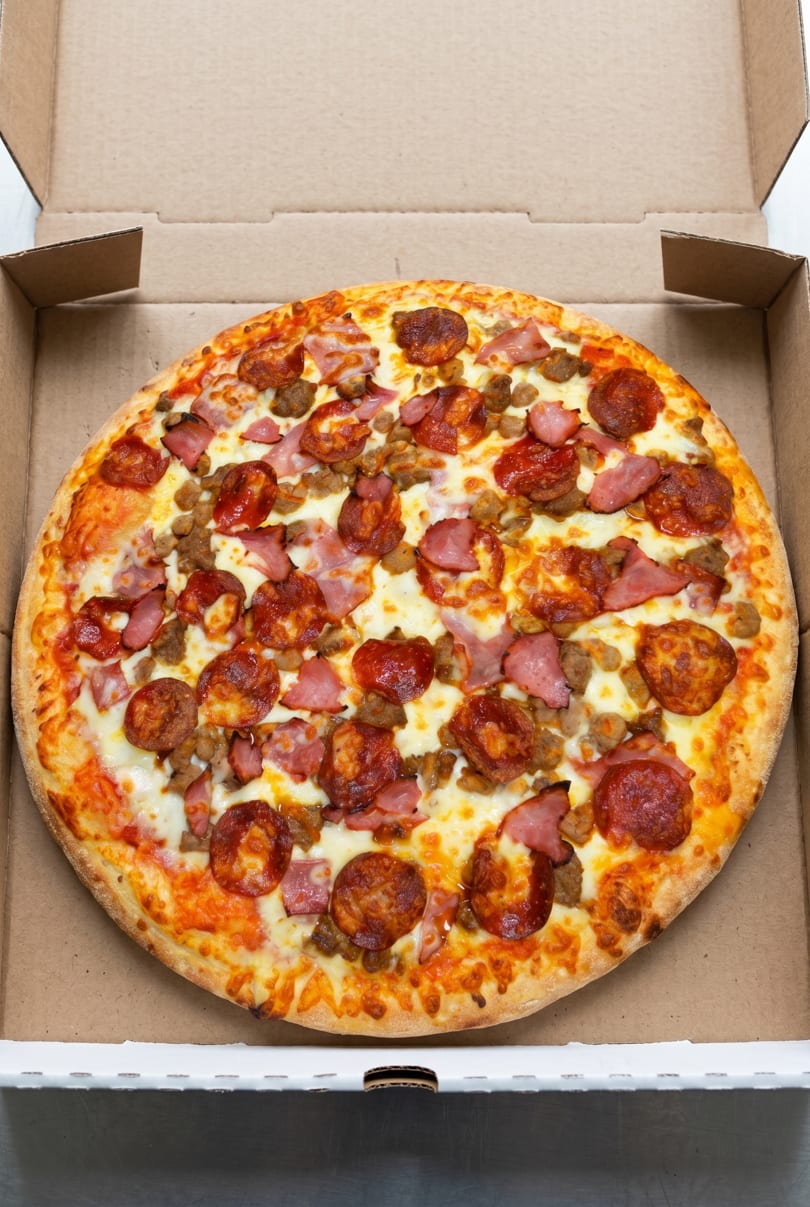A freshly baked meat lover's pizza with pepperoni, ham, and sausage in a cardboard box.